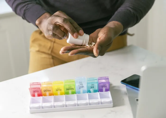 Close-up shot of an unrecognizable black man placing his prescription medication and nutritional supplements in a colorful daily pill box organizer. The pill organizer is sitting on the counter in the man's kitchen next to his cell phone and laptop comput